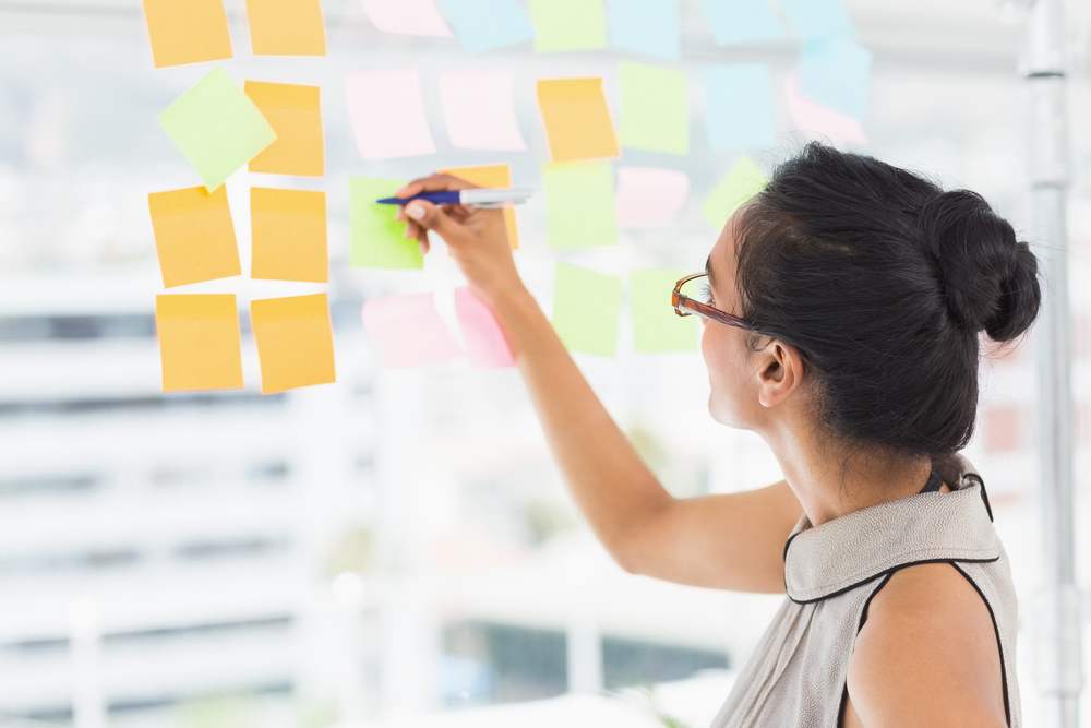 Smiling designer writing on sticky notes on window in creative office